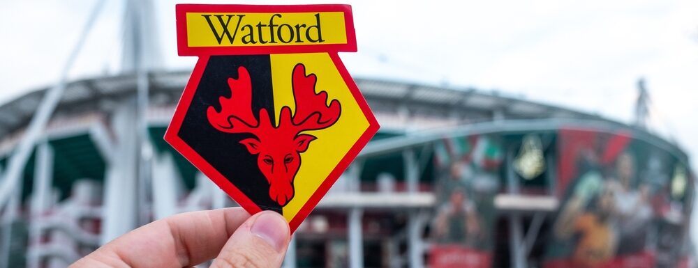 An image of the Watford Football Club badge, held up in front of Vicarage Lane football stadium.