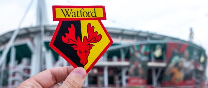 An image of the Watford Football Club badge, held up in front of Vicarage Lane football stadium.